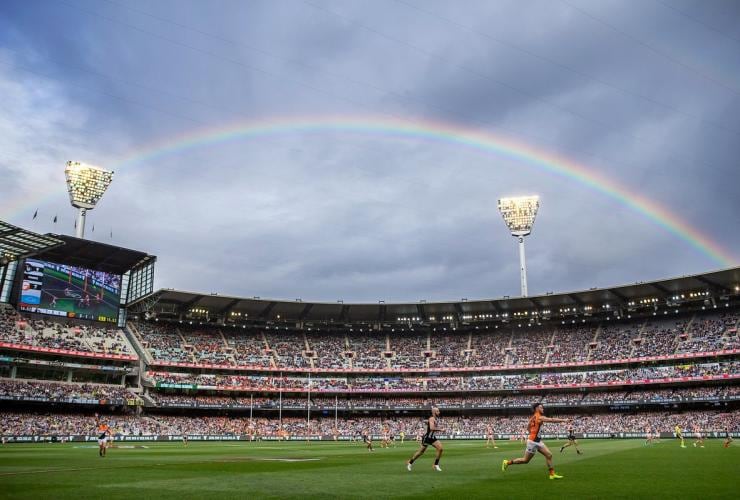 A field with people playing sport in the foreground in a stadium filled with spectators beneath a rainbow during Australian Rules Football at the Melbourne Cricket Ground, Melbourne, Victoria © Melbourne Cricket Ground