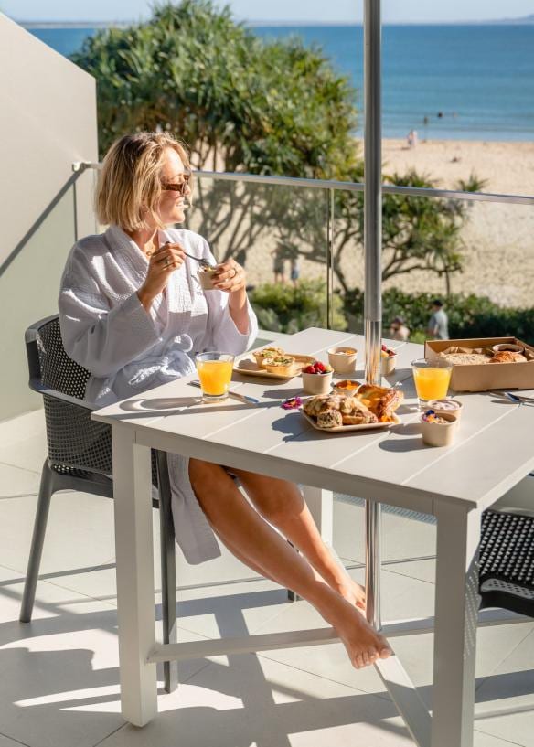 A person seated at a table with pastries and orange juice, wearing a robe and sunglasses while admiring the beach view at Noosa Heads, Sunshine Coast, Queensland © Tourism Australia
