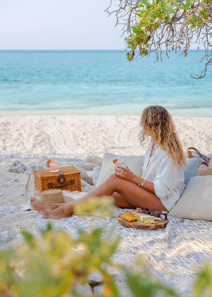 A person sitting on a picnic blanket with pillows, a picnic basket, a cheese board and two cocktails on a white sand beach near the light blue ocean on Wilson Island, Capricorn Coast, Queensland © Tourism Australia