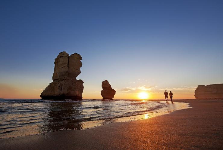 Two people walking along a beach during a bright sunset beside towering sandstone stacks standing in the ocean at Gibson Steps, Great Ocean Road, Victoria © Visit Victoria
