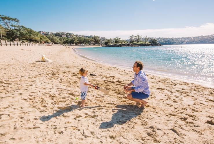 A person crouching down on sand by the ocean to play cricket with their child at Balmoral Beach, Sydney, New South Wales © Tourism Australia