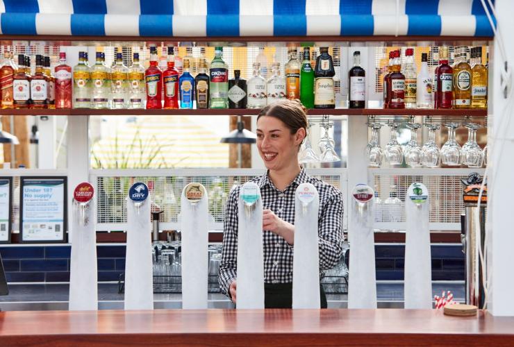 A smiling bar staffer stands behind beer taps, pulling a beer. Above her is a row of alcohol used to make cocktails, Portarlington Grand Hotel, Geelong and the Bellarine, Victoria © Tourism Australia