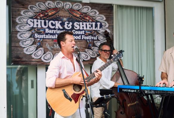 In front of a large banner showing the branding for the Shuck & Shell Seafood Festival are three musicians, playing instruments. They’re laidback and happy as they play live music to the festival crowd, Barrington Coast, New South Wales © Shuck & Shell Seafood Festival