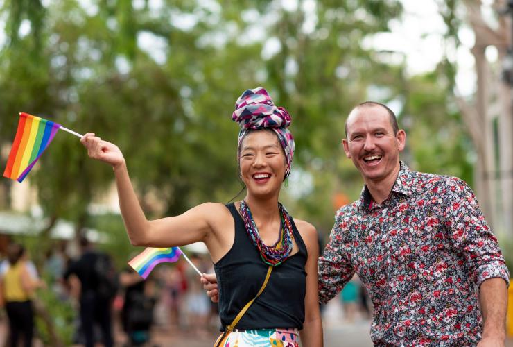 Two visitors smiling while waving pride flags at an outdoor celebration, FabAlice, Alice Springs, Northern territory © Tourism and Events NT/Helen Orr
