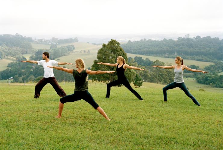 A group of people standing in a yoga pose with their arms outstretched on a grassy hill overlooking the Byron Bay hinterland at Gaia Retreat and Spa, New South Wales © Tourism Australia