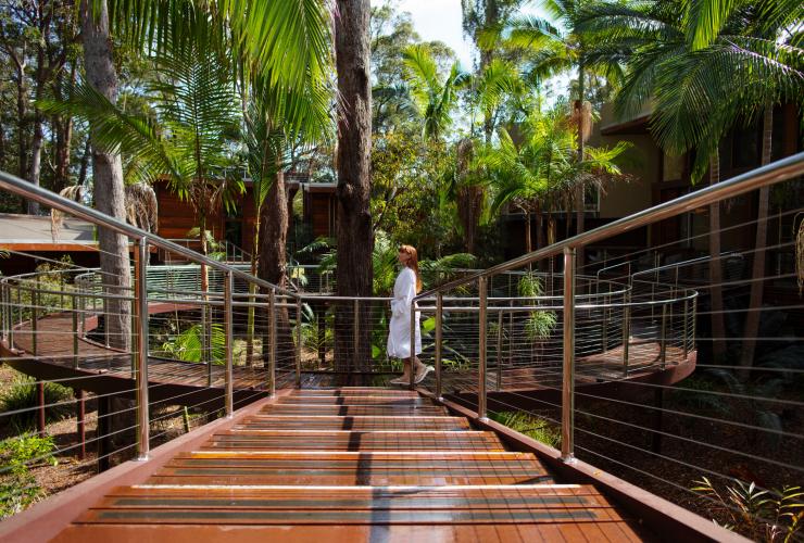 A woman wearing a white robe walking along an elevated wooden pathway surrounded by trees with a wooden building behind her at Gwinganna Lifestyle Retreat, Gold Coast, Queensland © Destination Gold Coast
