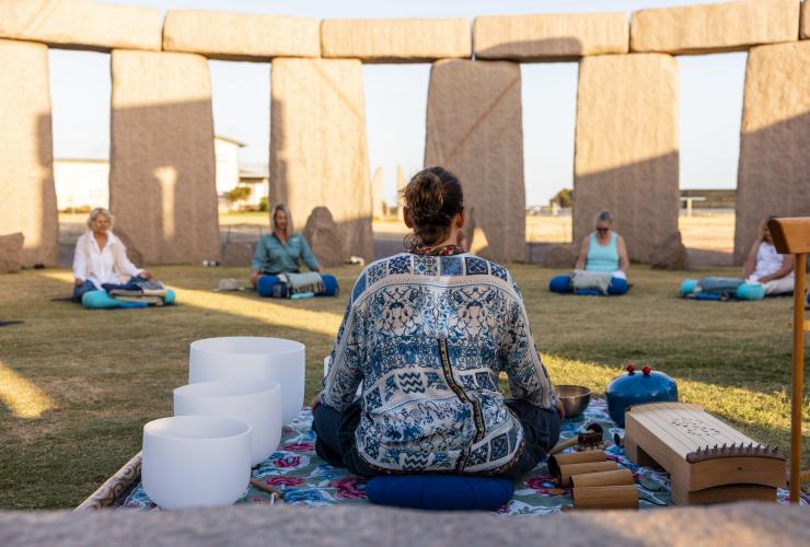 A yoga instructor seated among a variety of instruments with her back to the camera as a group of people sit spread out in yoga poses in front of her with Untamed Escapes, Esperance, Western Australia © Tourism Australia