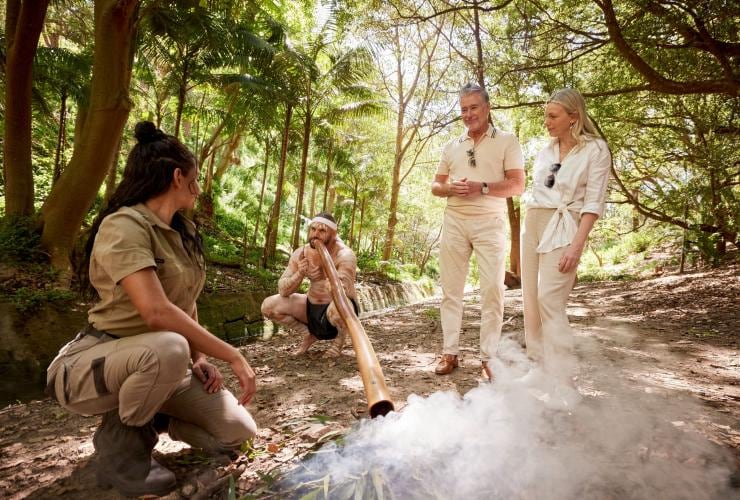 Two Aboriginal guides standing with two guests in bushland during a smoking ceremony and didgeridoo performance, Splendour Tailored Tours, Sydney, New South Wales © Tourism Australia