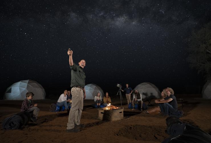 Tour guide pointing towards the night sky filled with stars while guests sit in a circle around a campfire listening with dome tents in the background at Earth Sanctuary, East MacDonnell Ranges, Northern Territory © Tourism Australia