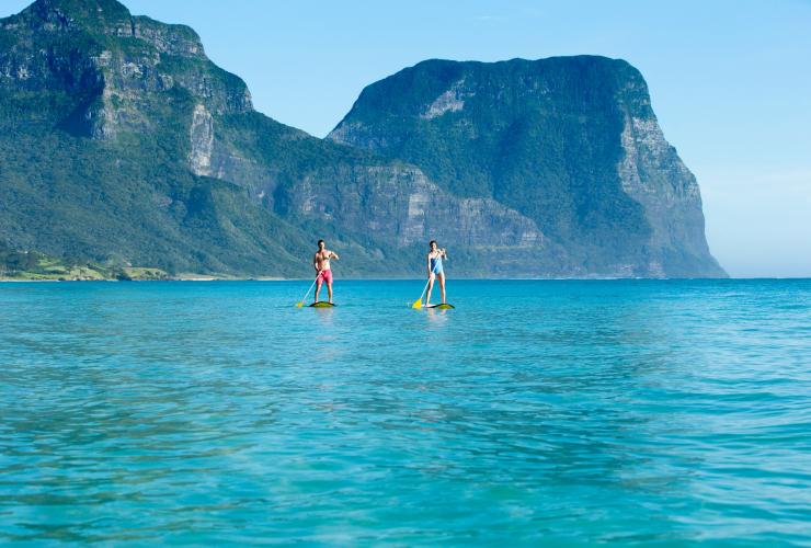 Man and woman stand-up paddle boarding on pristine blue water with Lord Howe Island in the background, NSW © Destination NSW