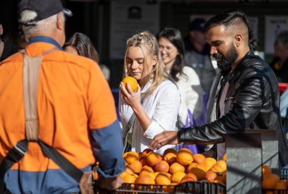 Illuminated by bright, morning sunshine, a lady is pictured smelling an orange she has picked up from a farm market stall at Capital Region Farmers Market, Canberra, Australian Capital Territory © Tourism Australia