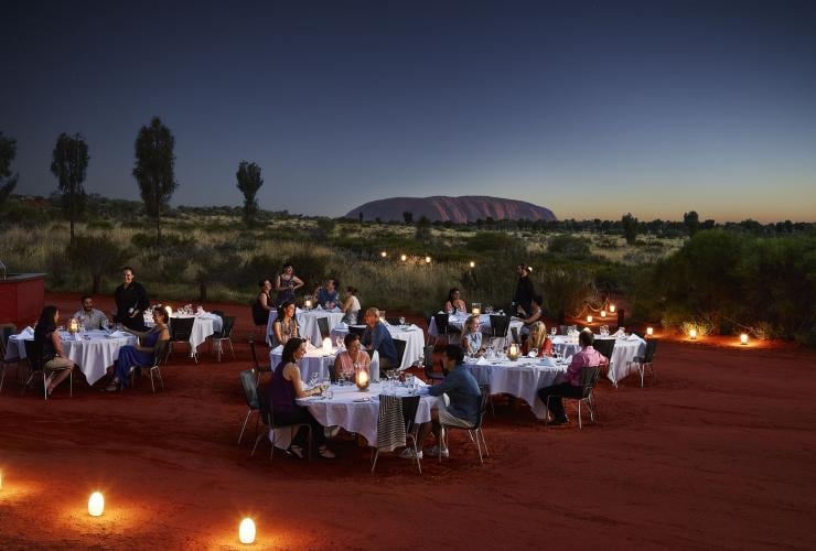 Several groups of people dining at tables on the red dirt with Uluru in the background at dusk at Sounds of Silence, Uluru, Northern Territory © Voyages