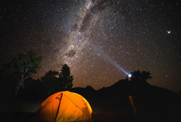 A man standing with his headlight pointing towards the starry night sky, beside a tent glowing from lights inside at the Dark Sky Park, Warrumbungle National Park, New South Wales © Destination NSW