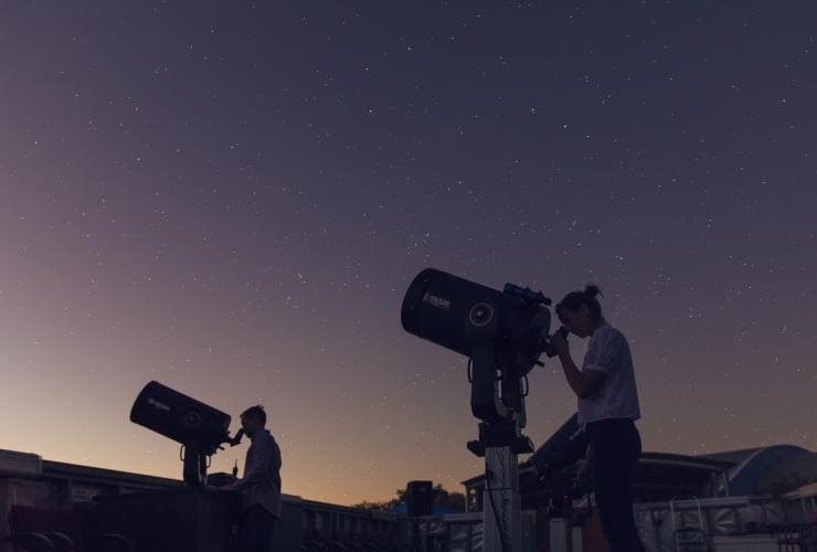 Two people looking through telescopes just after dusk with stars in the sky above them and the sun’s glow visible on the horizon at Cosmos Centre, Charleville, Queensland © Tourism and Events Queensland