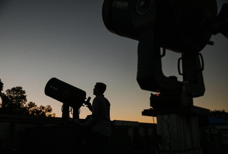 The sun setting over the horizon with a dark silhouette in the foreground of a person looking through a telescope towards the sky at the Cosmos Centre, Charleville, Queensland © Tourism and Events Queensland