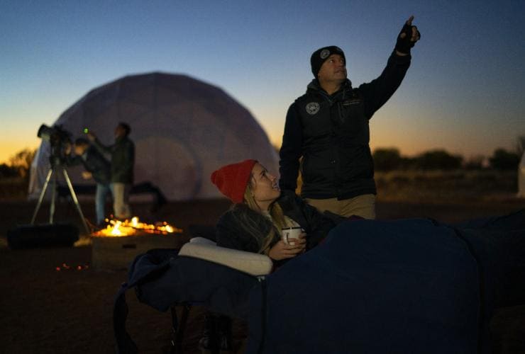 Tour guide pointing towards the sky at dusk beside a woman listening while sitting under a blanket with people looking through a telescope in the background at Earth Sanctuary, East MacDonnell Ranges, Northern Territory © Tourism Australia