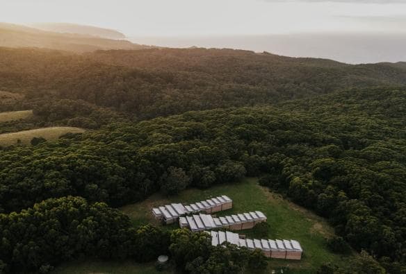 Aerial view over three rows of small buildings surrounded by greenery with the ocean visible in the distance at Alkina Lodge, Wattle Hill, Victoria © Alkina Lodge
