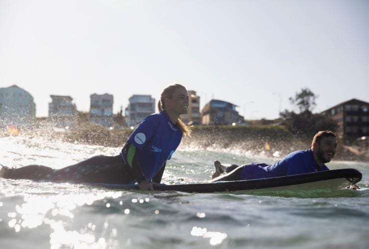 Two people laying on surfboards, smiling as they paddle away from the shore of a city beach, Lets Go Surfing, Bondi Beach, New South Wales © Destination NSW