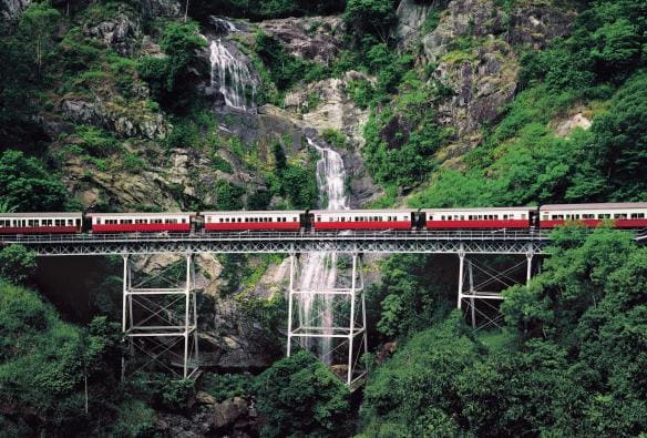 A train travelling on an elevated track past a large waterfall flowing through rainforest, Kuranda Scenic Railway, Kuranda, Queensland © Tourism and Events Queensland
