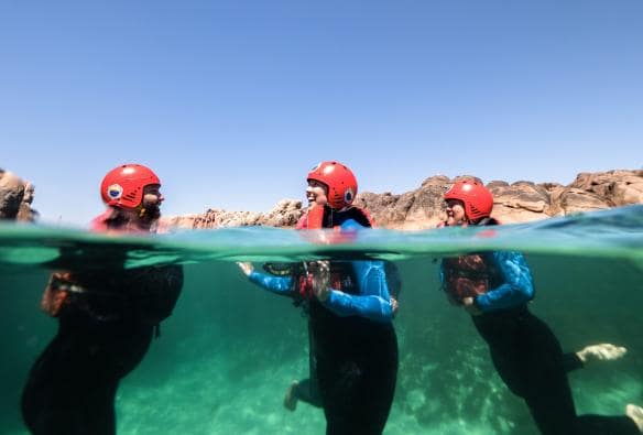 A semi-submerged shot shows a group of people wearing wetsuits, life vests and helmets, floating in turquoise ocean water with Margaret River Adventure Co, Margaret River, Western Australia © Tourism Australia