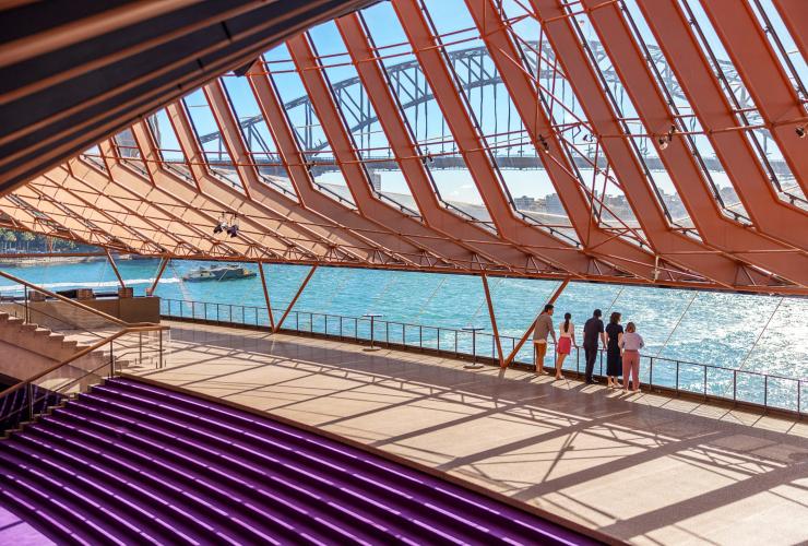 A tour group stands inside a large hall looking out onto the blue water and arched bridge of Sydney Harbour, Sydney, New South Wales © Tourism Australia