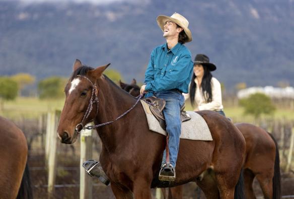Young man laughs as he rides a brown horse along the edge of a vineyard with Hunter Valley Horses, Hunter Valley, New South Wales © Tourism Australia