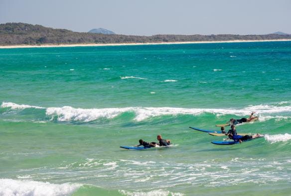 Several surfers lay on surfboards as a white-capped wave rolls in behind them at Noosa Main Beach, Noosa, Queensland © Tourism Australia