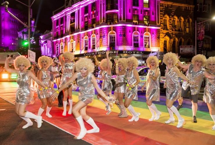 A group of people wearing blonde wigs and silver dresses dancing across a road painted with a rainbow, Sydney Mardi Gras, Sydney, New South Wales ©   James Horan/Destination NSW