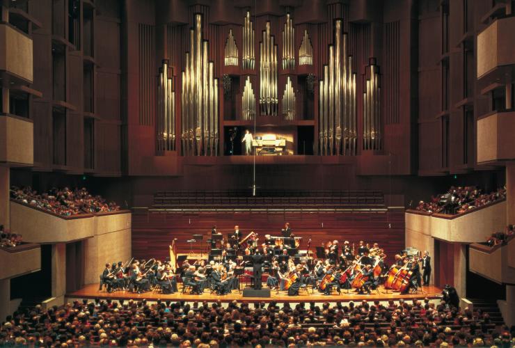 An orchestra on stage in a wood-panelled theatre performing in front of a crowd at Queensland Performing Arts Centre, Brisbane, Queensland © Tourism and Events Queensland
