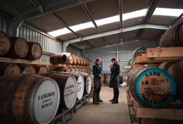 Two women standing between rows of barrels while tasting spirits at Lark Distillery, Hobart, Tasmania © Tourism Australia