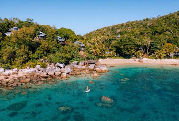 View over the clear blue ocean towards a person paddle boarding close to a rocky headland beside a white sand beach, with villas tucked among lush greenery at Bedarra Island Resort, Great Barrier Reef, Queensland © James Vodicka