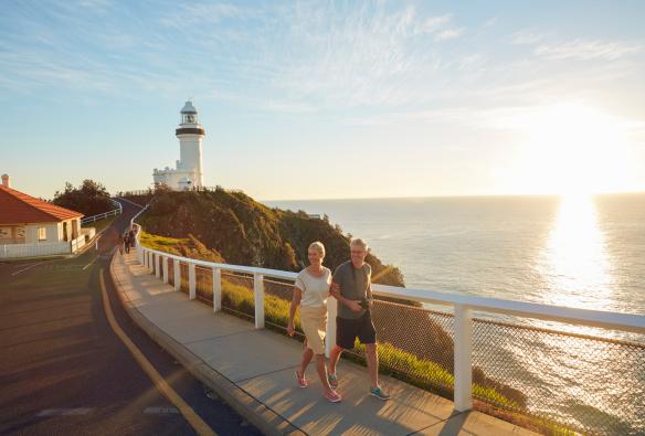 A couple walking along a pathway beside the ocean with Cape Byron Bay Lighthouse behind them, Byron Bay, New South Wales © Tourism Australia