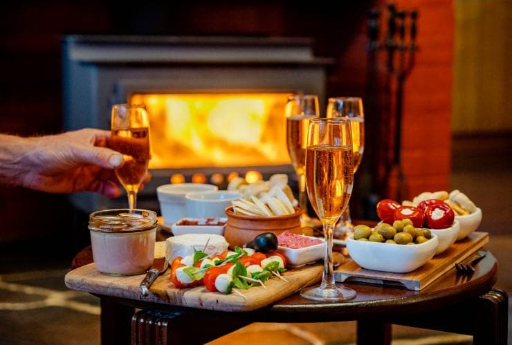 A hand reaching for a glass of Champagne, which is set among platters of olives and cheese with a fireplace roaring in the background on the Southern Ocean Walk in South Australia © Tourism Australia