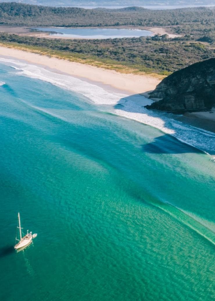 Aerial view over a boat in turquoise water near a white sand beach surrounded by bushland.
