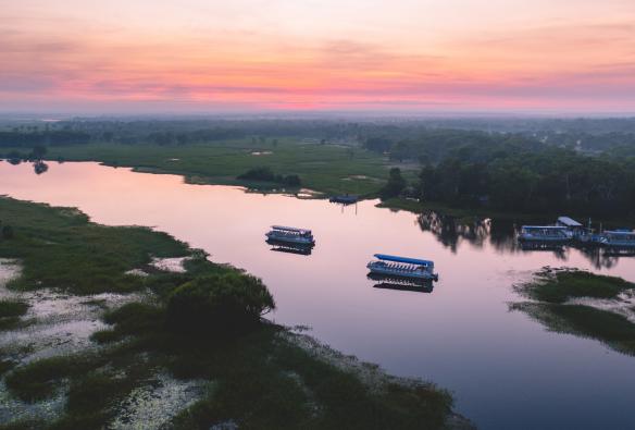 Vue aérienne de bateaux qui naviguent sur une rivière calme, entourée de verdure, reflétant les teintes rose et orange pastel du ciel au coucher du soleil à Yellow Water Billabong, Kakadu National Park, Territoire du Nord © Tourism NT/Salty Wings