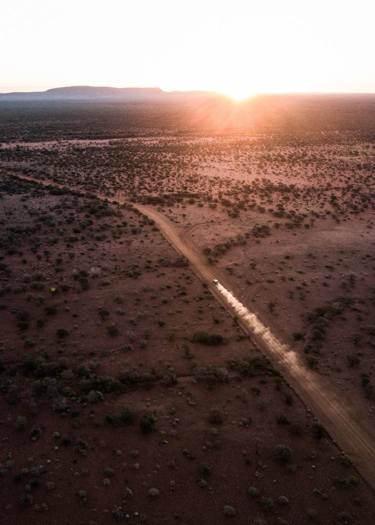 Vue aérienne d'une voiture soulevant de la poussière sur un chemin de terre dans l'arrière-pays, se dirigeant vers une formation rocheuse au loin à Mount Augustus, Wheatbelt, Australie Occidentale © Australia's Golden Outback