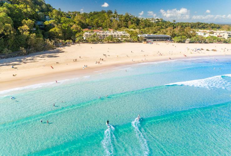 Vue aérienne de l'eau bleu vif et du sable aux reflets dorés, tandis que des surfeurs chevauchent les vagues vers le rivage à Noosa Main Beach, Noosa, Queensland © Tourism and Events Queensland