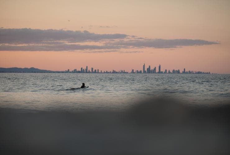 Un surfeur est allongé sur sa planche de surf dans l'océan, les bâtiments du paysage urbain de la Gold Coast se dessinant derrière, à Snapper Rocks, Gold Coast, Queensland © Tourism and Events Queensland