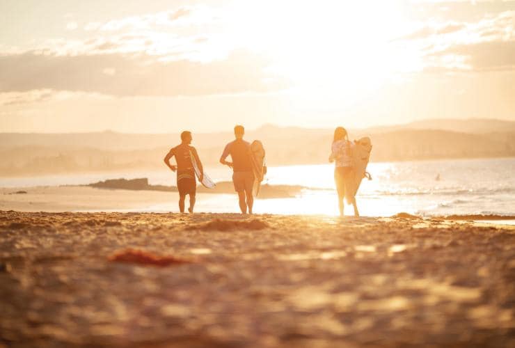 Trois surfeurs face au lever du soleil courant sur le sable en direction de l'océan à Snapper Rocks, Gold Coast, Queensland © Tourism and Events Queensland