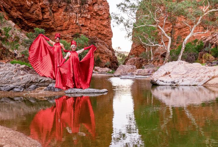 Deux drag queens vêtues de robes rouges spectaculaires posant à côté d'un point d'eau de l'outback entouré de falaises de roche rouge, FabAlice, Alice Springs, Territoire du Nord © Tourism and Events NT/Helen Orr 