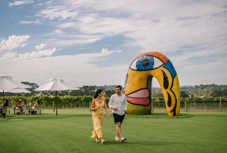 Deux personnes traversent une pelouse sous leurs parapluies, avec en arrière-plan un vignoble et une grande sculpture colorée au Pt. Leo Estate, Mornington Peninsula, Victoria © Tourism Australia
