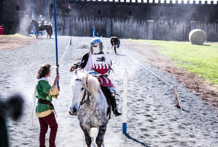Cavalier à cheval déguisé en chevalier tandis qu'une autre personne habillée en costume d'époque lui tend une épée à Kryal Castle, Ballarat, Victoria © Kryal Castle