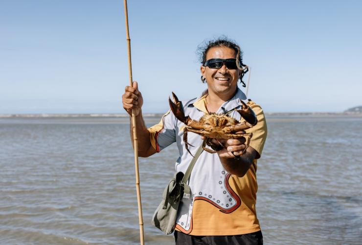 Un guide touristique aborigène debout dans des eaux peu profondes et tenant dans ses mains une lance et un crabe des palétuviers avec Walkabout Cultural Adventures, Port Douglas, nord tropical du Queensland, Queensland © Tourism Australia