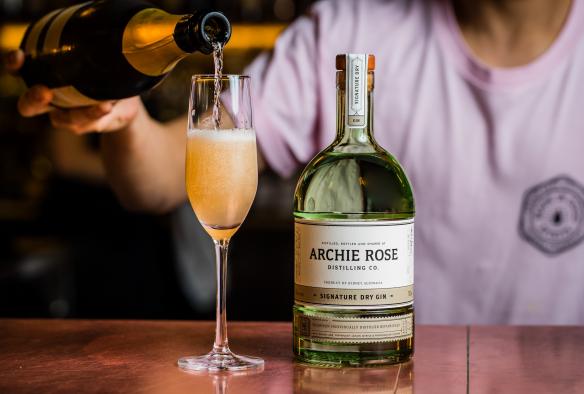Gros plan d'un barman versant du champagne dans une flûte, posée à côté d'une bouteille de « Signature Dry Gin » à Archie Rose, Sydney, Nouvelle-Galles du Sud © Archie Rose