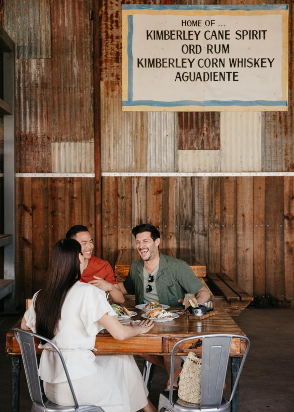 Trois amis assis autour d'une table sur laquelle sont posées des assiettes de nourriture au Hoochery Distillery Café, Kununurra, Australie Occidentale © Tourism Western Australia