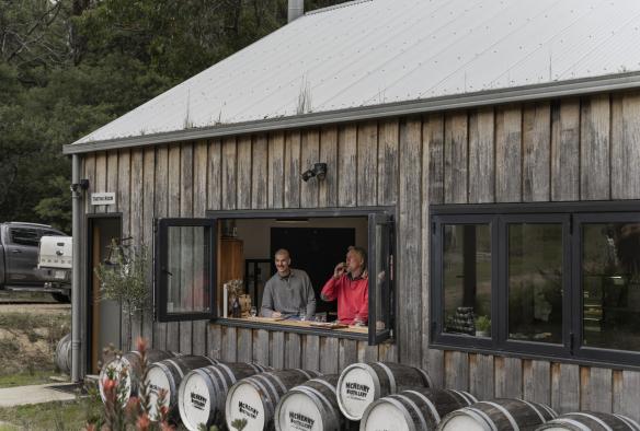 Un petit bâtiment rustique dans lequel se trouvent deux hommes, debout près de la fenêtre, regardant à l'extérieur tout en sirotant des boissons à la McHenry Distillery, Tasman Peninsula, Tasmanie © Tourism Australia