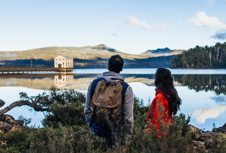 Couple en pleine randonnée et faisant une pause sur un belvédère du Lake St Clair avec vue sur Pumphouse Point au loin, Lake St Clair, Tasmanie © Adam Gibson