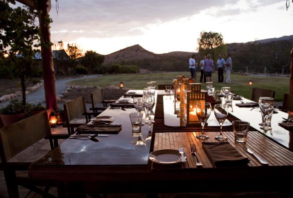 Une élégante table dressée pour le dîner, dont l'effet miroir est mis en valeur au premier plan par la lumière des bougies, avec un groupe de personnes buvant du vin sur la pelouse au loin au Arkaba Conservancy, Flinders Ranges, Australie du Sud © Arkaba Conservancy/Randy Larcombe
