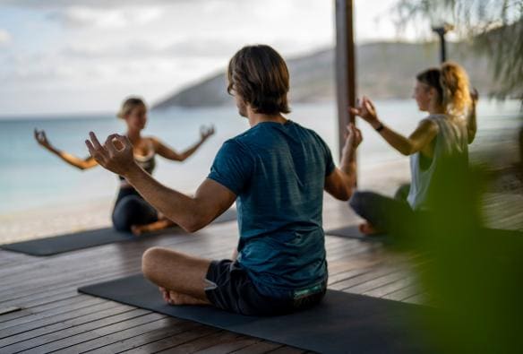 Un couple assis sous une cabane sur la plage faisant une posture de yoga en imitant leur professeure au Lizard Island Resort, Lizard Island, Queensland © Tourism and Events Queensland
