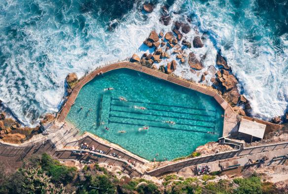 Vue aérienne de personnes nageant dans l'eau turquoise des Bronte Baths, avec les vagues de l'océan s'écrasant contre les rochers près de la piscine, Bronte Beach, Sydney, Nouvelle-Galles du Sud © Tourism Australia
